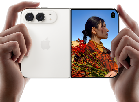 Rozevřený nebesky bílý iPhone Duo, dvě ruce ho drží na šířku, na levém panelu je vidět dvojitá 48Mpx Fusion zadní fotosoustava a vpravo se na vnějším displeji zobrazuje osoba, která se nechává fotit a díky funkci Duo Preview vidí při focení svůj obličej