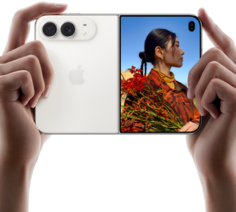 Rozevřený nebesky bílý iPhone Duo, dvě ruce ho drží na šířku, na levém panelu je vidět dvojitá 48Mpx Fusion zadní fotosoustava a vpravo se na vnějším displeji zobrazuje osoba, která se nechává fotit a díky funkci Duo Preview vidí při focení svůj obličej