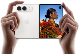 Rozevřený nebesky bílý iPhone Duo, dvě ruce ho drží na šířku, na levém panelu je vidět dvojitá 48Mpx Fusion zadní fotosoustava a vpravo se na vnějším displeji zobrazuje osoba, která se nechává fotit a díky funkci Duo Preview vidí při focení svůj obličej