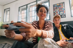Eine junge Frau sitzt auf einer Couch, hält ein Gamepad in beiden Händen und schaut amüsiert