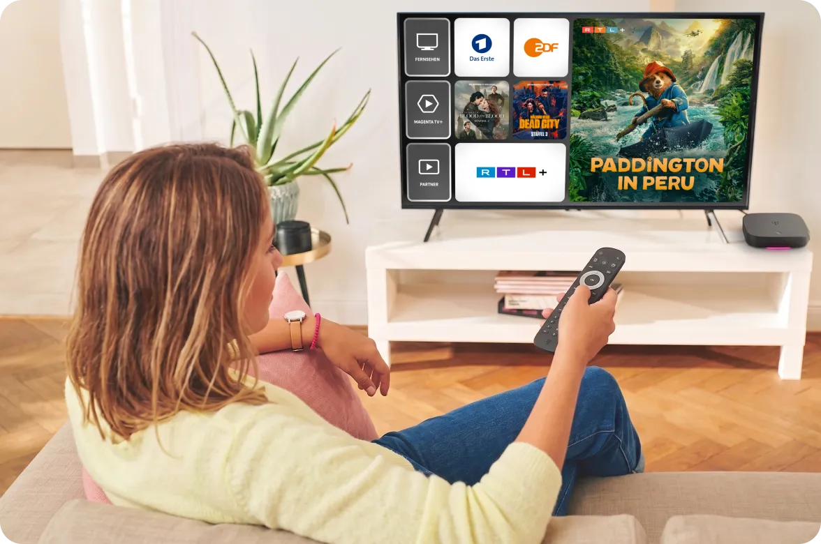 Frau mit Fernbedienung auf dem Sofa vor einem Fernseher mit der MagentaTV Oberfläche: Das Erste, ZDF und RTL+ sowie der Film Paddington in Peru