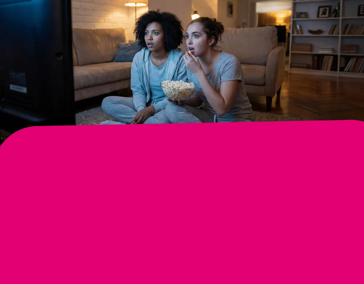 Zwei junge Frauen sitzen abends in einem gemütlichen Wohnzimmer auf dem Boden, essen Popcorn und schauen gespannt in Richtung eines Fernsehers.