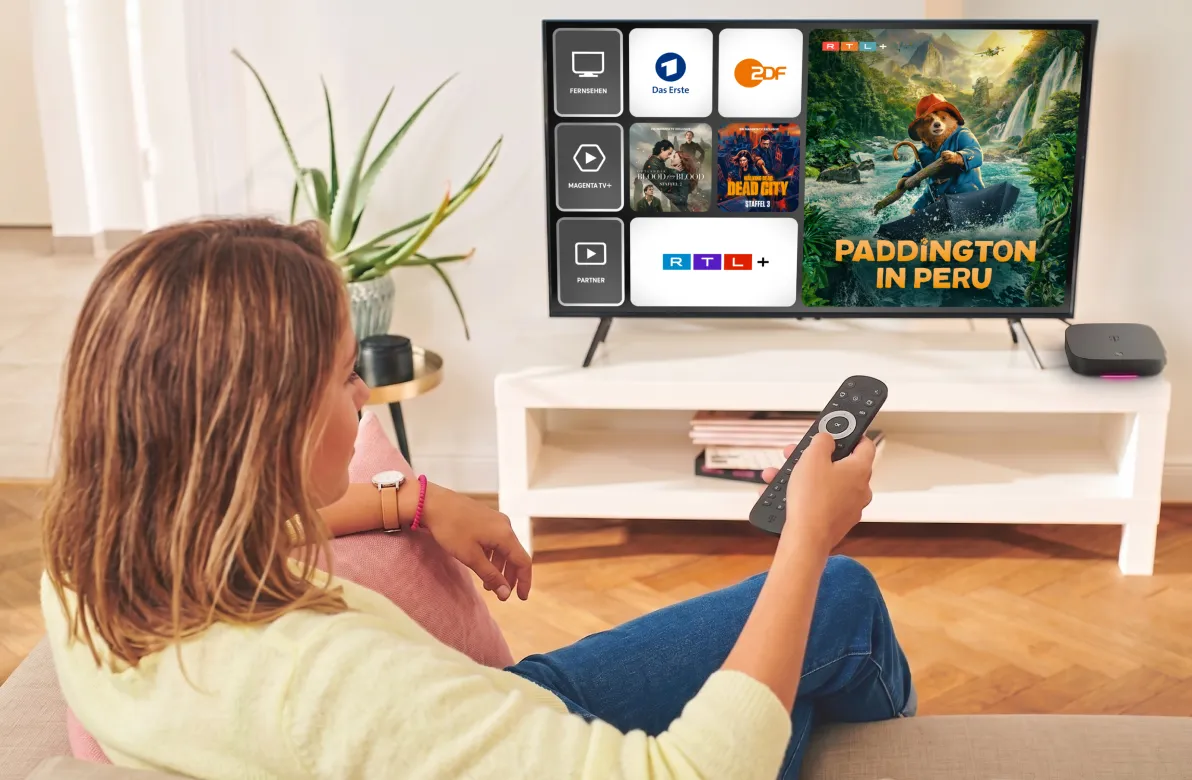 Frau mit Fernbedienung auf dem Sofa vor einem Fernseher mit der MagentaTV Oberfläche: Das Erste, ZDF und RTL+ sowie der Film Paddington in Peru