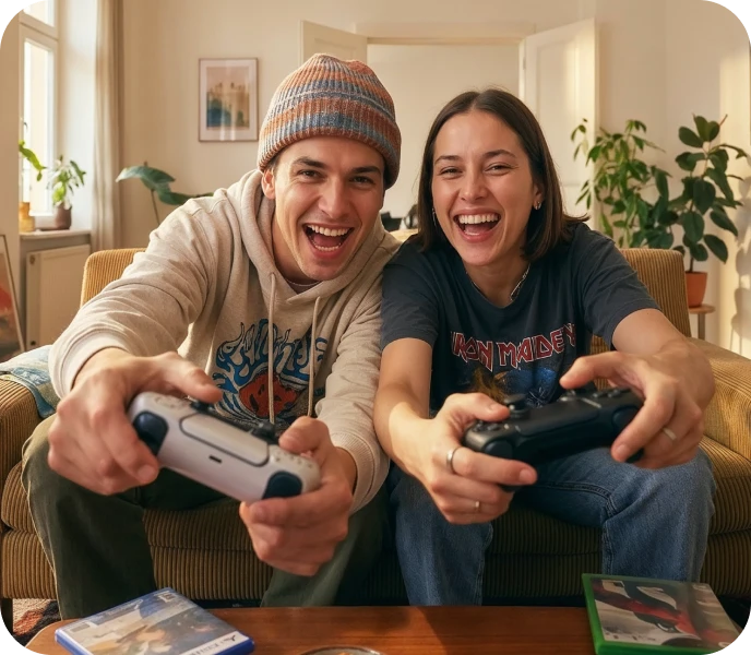 Zwei junge Menschen sitzen lachend auf einem Sofa und spielen gemeinsam Videospiele mit Controllern.