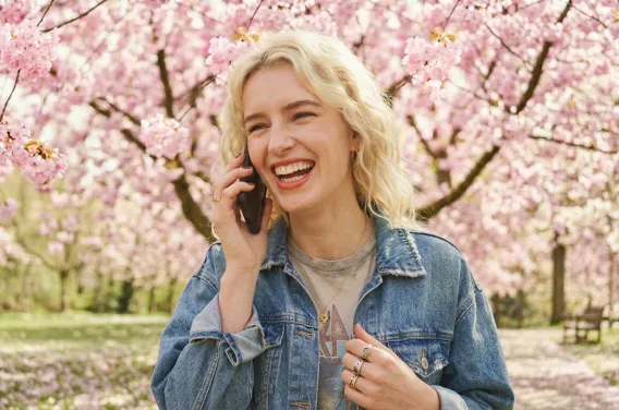 Blonde Frau in Jeansjacke steht unter blühenden Kirschbäumen und telefoniert