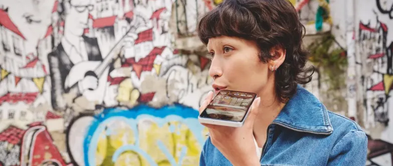 Eine Frau steht an einer Wand, die mit Graffiti gestaltet ist und nutzt die Sprachfunktion auf ihrem Smartphone.