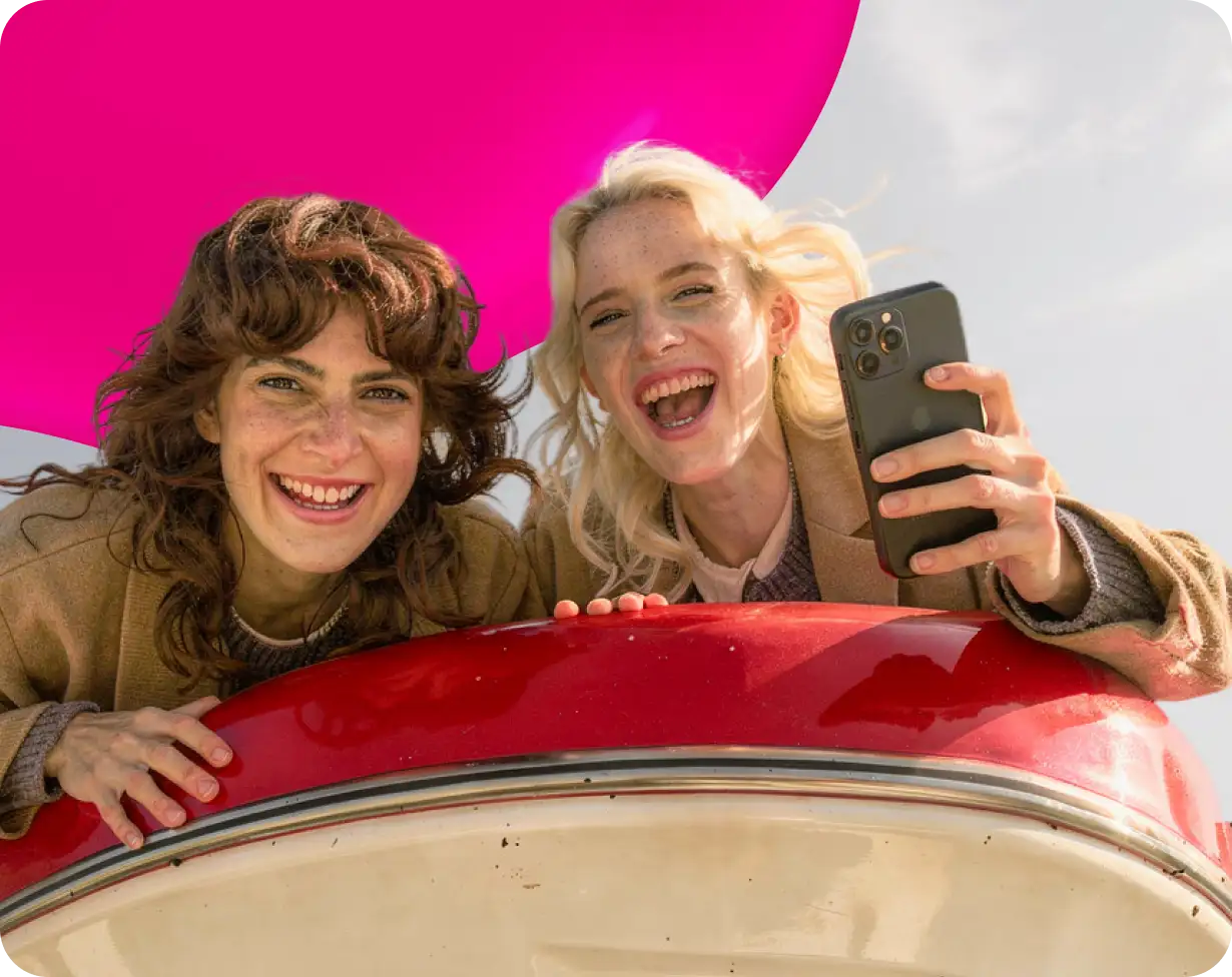Zwei lachende Frauen lehnen über das Dach eines roten Autos, eine hält ein Smartphone in der Hand.