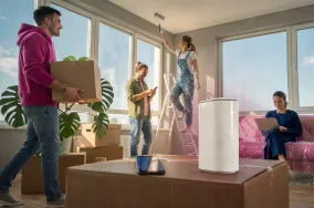 Junge Leute beim Einzug in eine neue Wohnung – ein Mann trägt einen Umzugskarton, während Freunde beim Auspacken helfen. Im Vordergrund steht ein weißer WLAN-Router.