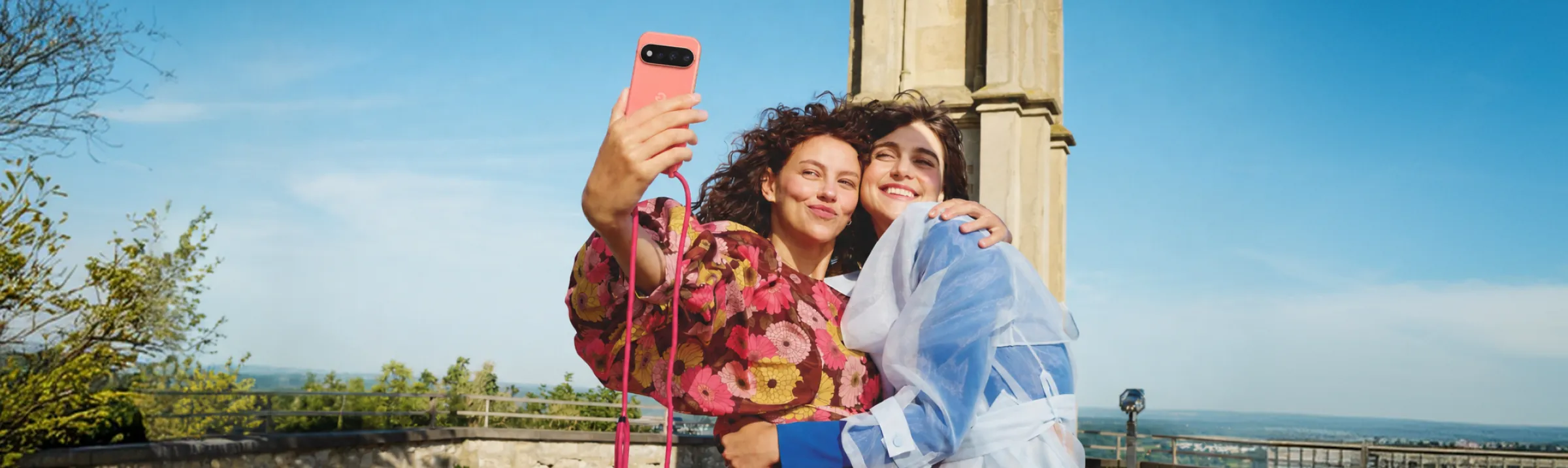 Zwei junge Frauen umarmen sich und machen lächelnd ein Selfie mit einem Smartphone an einer Handykette