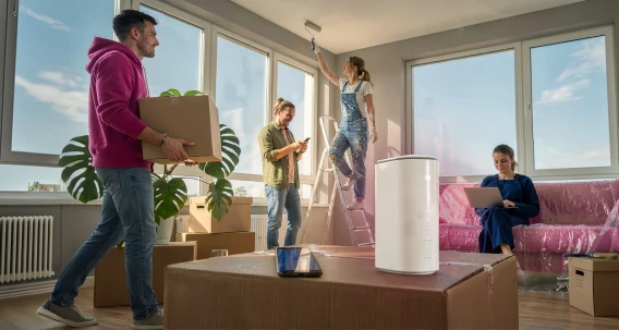 Junge Leute beim Einzug in eine neue Wohnung – ein Mann trägt einen Umzugskarton, während Freunde beim Auspacken helfen. Im Vordergrund steht ein weißer WLAN-Router.