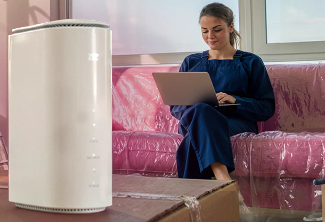 Eine im Vordergrund stehende Speedbox auf einem Umzugskarton, dahinter eine auf einem in Schutzfolie gehüllten Sofa sitzende Frau mit einem Laptop auf dem Schoß